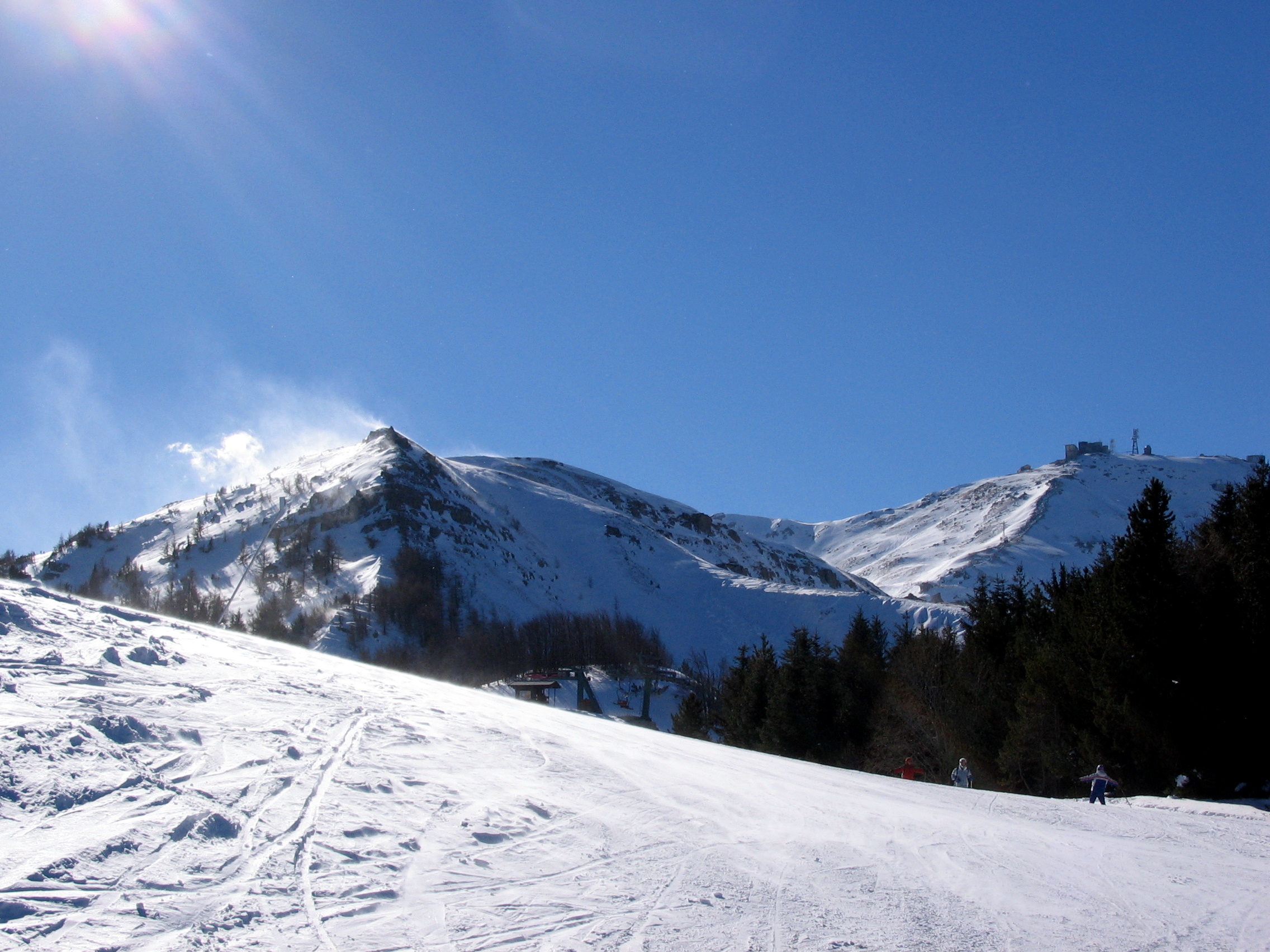 Le creste innevate del Monte Cimone sotto un cielo blu, con una pista da sci in primo piano e i boschi di abeti a destra